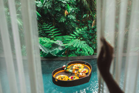 Floating breakfast in jungle swimming pool, tropical resort. Black rattan tray in heart shape, Valentines day or honeymoon surprise, view from above. Exotic summer diet. Tropical beach lifestyle.の写真素材