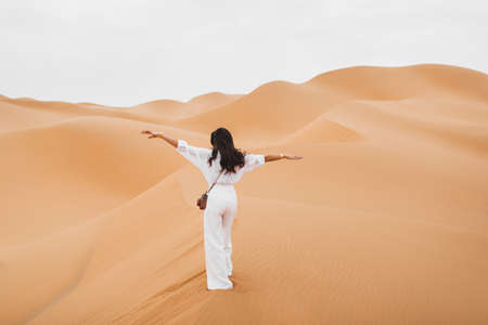 Woman in stylish white shirt and pants with retro photo camera happy to explore Sahara desert sand dunes in Morocco. View from the back.の写真素材