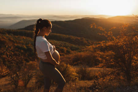 Young pregnant woman walking outdoors with amazing autumn mountain and forest view. Warm sunlight. Wellness and vitality in maternity time.の写真素材