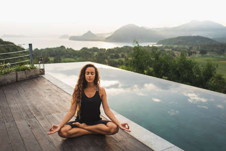 Woman practicing yoga in lotus position on infinity poolside with beautiful ocean and mountain view in morning. Healthy lifestyle, spiritual and emotional concept. Awakening.の写真素材