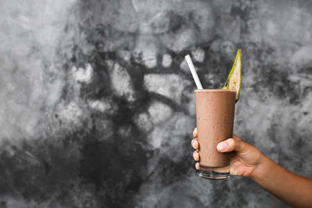 Glass of fresh cold chocolate milk shake in woman hands on background of gray concrete cement textured wall. Cocoa smoothie with banana piece as decoration. Summer concept.の写真素材