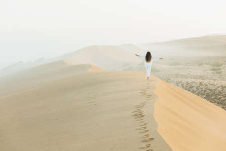 Woman walking on top of huge sand dune in Morocco Sahara desert. Beautiful warm sun light and mist in morning. View from behind. Freedom concept.の写真素材