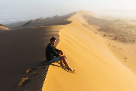 Man enjoying sunrise on top of huge sand dune. Beautiful warm sun light. Sahara desert, Morocco.の写真素材