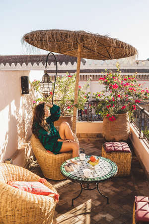 Woman enjoying breakfast on beautiful lounge hotel terrace in downtown of Marrakech at sunrise. Moroccan design with vintage elements - metal lantern, wicker chair, round table, and umbrella.の写真素材