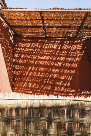 Background of light and shadow on red terracotta wall in Morocco. Abstract geometric texture, straw wooden elements.の写真素材