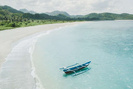 Old blue wooden traditional fishing boat on turquoise ocean surface in Indonesia. Amazing white sand beach coastline. Aerial drone shot from above.の写真素材