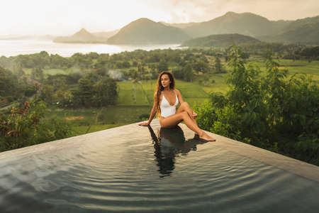 Dream journey in Asia. Woman sitting on edge luxury infinity pool an enjoying amazing mountain, rice...