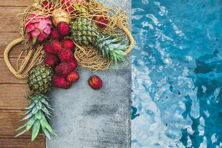 Flat lay of fresh tropical fruits pineapple, rambutan, dragon fruit in reusable mesh bag on pool...