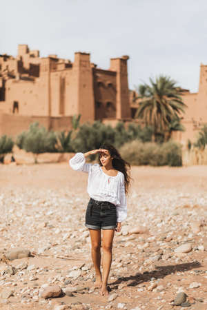 Young curly woman eastern appearance walking on background of kasbah Ait-Ben-Haddou. Travel in Morocco, Ouarzazate. Summer vacations, travel lifestyle concept.の写真素材