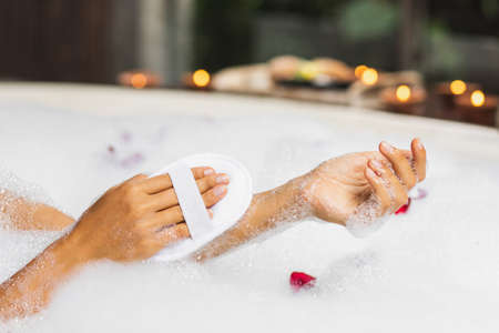 Woman washing hands in bath tub with foam bubbles and use natural jute sponge. Hands close-up. Spa...