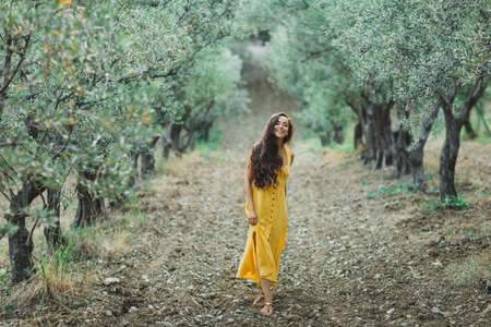 Young happy smiling woman walking in olive tree garden. Yellow linen summer dress. Trendy color and textile style.の写真素材