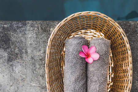 Two gray hand towels in wicker basket on poolside in hotel spa. Pink frangipani flower as decoration. Beauty treatment.の写真素材