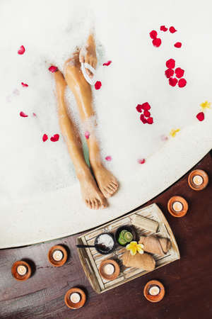 Woman legs in bath tub with foam bubbles and candles around. Romantic spa set with lime, scrub,...