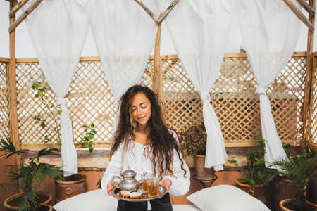 Beautiful young oriental woman holding tray with traditional moroccan mint tea, cookies and silver...