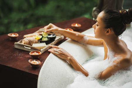 Woman taking a bath. Tub with foam bubbles. Romantic spa treatment, body care. Natural cosmetics set on wooden tray.の写真素材