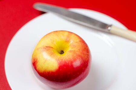 ripe red-yellow apple with a knife on a white plate against the red background