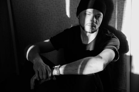 a sad young man in black sits on the floor leaning against the wall with his back. black and white.の写真素材