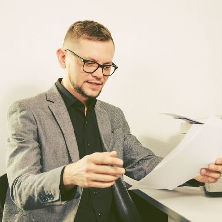 man with glasses and jacket smiles studying documents at table with laptop and printer at office. square view.の写真素材