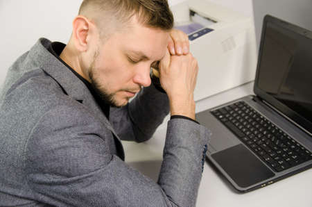 tired man with closed eyes and head leaning against hands at table with laptop in officeの写真素材