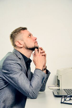 man with chin in hands thoughtfully looking upwards sitting at table with laptop in officeの写真素材