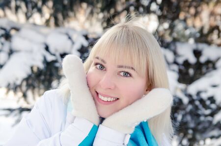 A beautiful blonde in sports clothes warms her cheeks with warm mittens in the forest against the background of snow-covered fir treeの写真素材