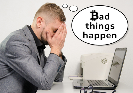 A sad man in a suit with his hands covering his face behind his desk with a laptop thinks that bad things happen. The symbol bitcoin is used. The concept of the collapse of bitcoin or the loss of savings of crypto currency.の写真素材