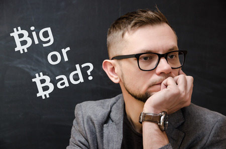 Man in a jacket and glasses on the background of a black school board on which it is written Big or Bad? The symbol bitcoin is used. The concept of thinking about the future bitcoin. The concept of thinking about the stability and reliability of bitcoin.の写真素材