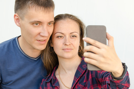 guy with short haircut in blue T-shirt and girl with dark long hair in red and blue checkered shirt take selfie on the phone. close view.の写真素材
