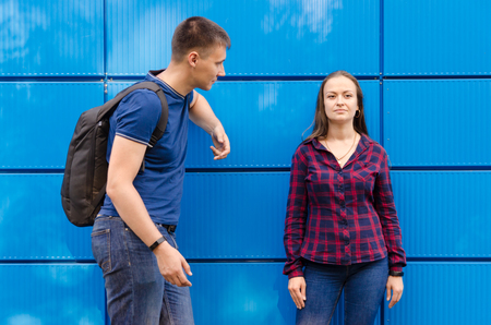 guy in blue T-shirt and backpack on his back look at girl dressed in blue jeans and plaid shirt against blue wall.の写真素材