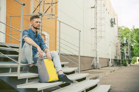 young man in jeans and a denim shirt is sitting on the stairs of an industrial building next to his...