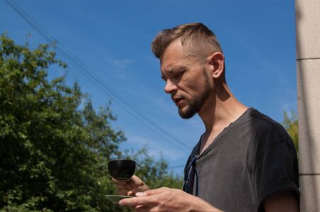 young bearded man with a cup of coffee in his hand against the blue sky on a sunny summer day.
