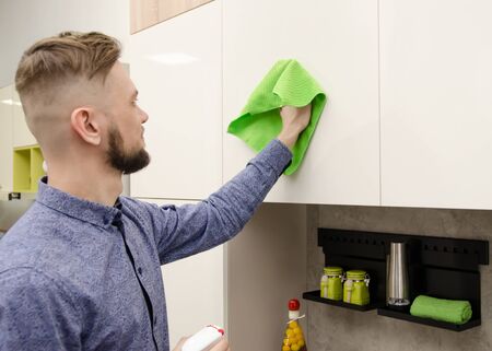 Attractive bearded man cleaning the handleless cabinet doors with a green rag in a modern kitchen.の写真素材