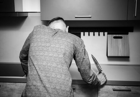 hunched man with a knife in his hand stands with his back to the camera in the kitchen.
