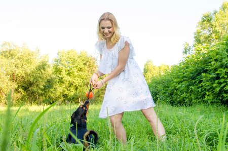 A beautiful blonde young woman in a light dress holds a ball in her hand for which a cute dog clung to.の写真素材