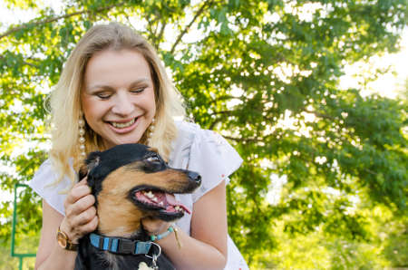 A beautiful smiling blonde young woman in a light dress with tenderness hugs her cute dog.の写真素材