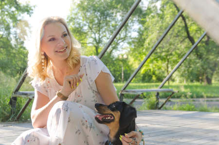 A beautiful blonde young woman in a light dress is playing with her cute dog on a summer evening.の写真素材
