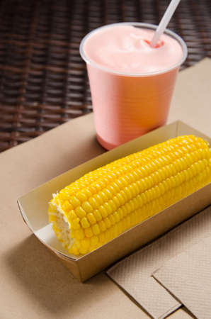 an ear of boiled corn in an eco-friendly cardboard box next to a milkshake.の写真素材