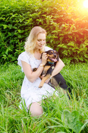 A beautiful blonde young woman sitting in green grass holds her cute dog in her arms and hugs her gently. Vertical view.の写真素材