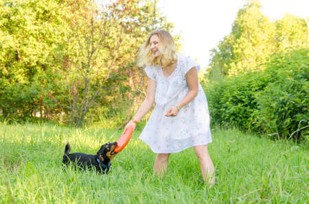 A blonde young woman in a light dress is holding a flying disc that a cute dog clung to.の写真素材