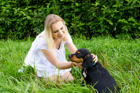 Beautiful blonde young woman sitting in green grass plays with her cute dog.の写真素材