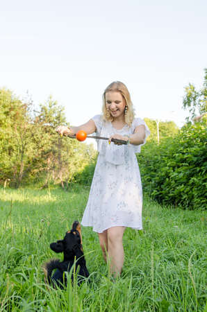 A beautiful blonde young woman in a light dress shows the ball to her cute dog to make her jump. Vertical view.の写真素材