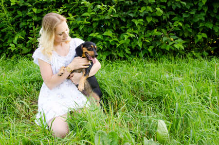 A beautiful blonde young woman sitting in green grass holds her cute dog in her arms.の写真素材
