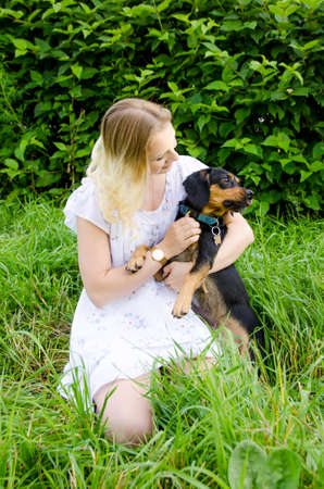 A beautiful blonde young woman sitting in green grass holds her cute dog in her arms. Vertical view.の写真素材