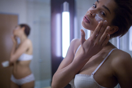 Beautiful young African woman spreading cream on her face and smiling while standing against a mirror in bathroomの写真素材