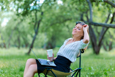 Girl using laptop on lunch break in city park and hold coffe in her handの写真素材