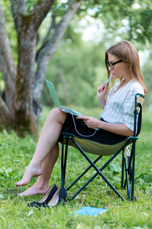 Young woman with a laptop sitting in the Park on a folding chair and holding a pen in her handの写真素材