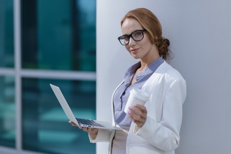 Young woman with a Cup of coffee in her hands looking at the laptop, standing next to her officeの写真素材