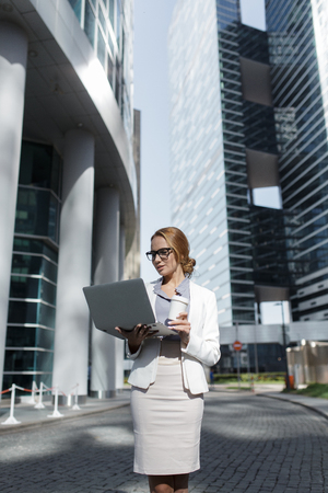 Young beautiful business woman with cup of coffee and laptop in her hands standing among the downtownの写真素材