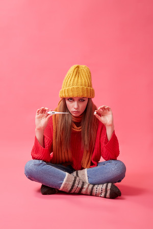 A sick woman in a yellow hat holds a medical thermometer and pills in her hands. Image on the pink backgroundの写真素材