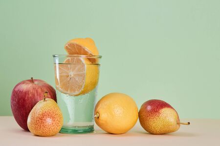 Glass with lemon water. Parts of juicy lemon. Red apple and pear. Summer healthy fruits. Copy space on green backgroundの写真素材